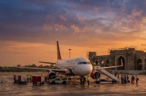 "A photorealistic, wide-angle shot of Darbhanga Airport bustling with activity during a golden sunset. In the foreground, a modern commercial aircraft is parked on the tarmac with passengers boarding. The background subtly features the architectural structure of the upcoming new terminal inspired by the historic Darbhanga Fort. The sky has faint, ethereal patterns of Madhubani art blending with the clouds, symbolizing the culture of Mithila. The image should evoke a sense of progress, hope, and vibrant energy. High definition, 8k resolution, cinematic lighting."